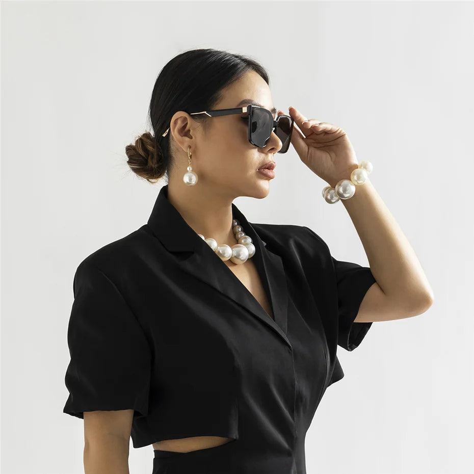 Une femme portant un blazer noir et des lunettes est posant, mettant en valeur un élégant collier imitation perles.