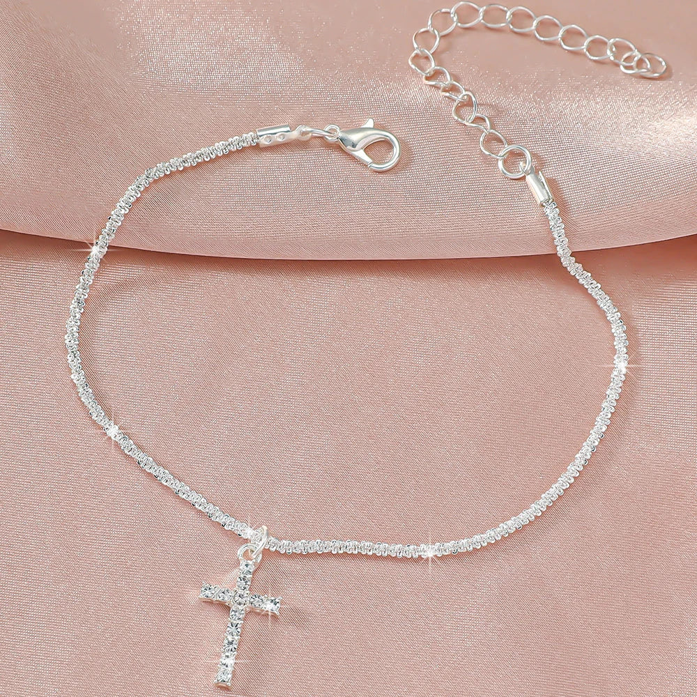 A delicate silver-plated chain anklet with a cross charm displayed on a pink fabric background.