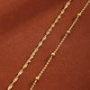 Two strands of gold plated beaded anklets laid diagonally on a reddish-brown fabric.