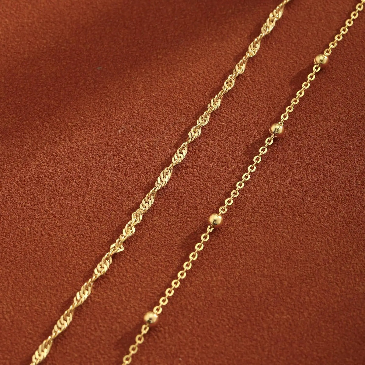 Two strands of gold plated beaded anklets laid diagonally on a reddish-brown fabric.