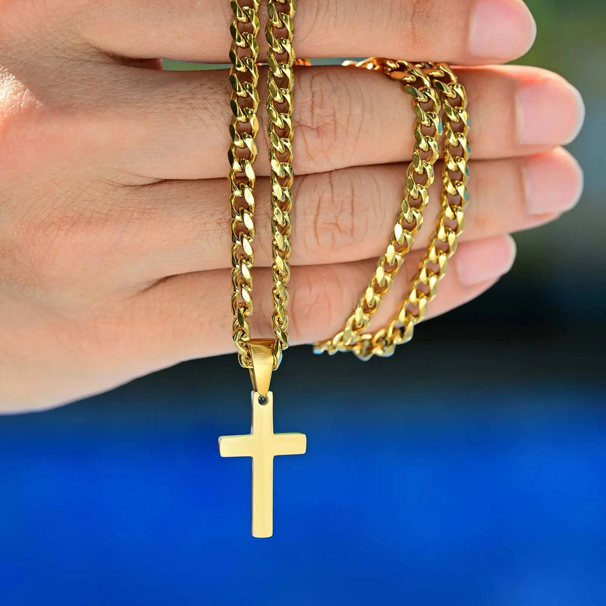 A hand holds a gold-colored stainless steel Cuban link necklace with a cross pendant.