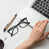 A hand holds a pair of black square anti-blue light eyeglasses next to a laptop keyboard and a wooden pen on a white desk.