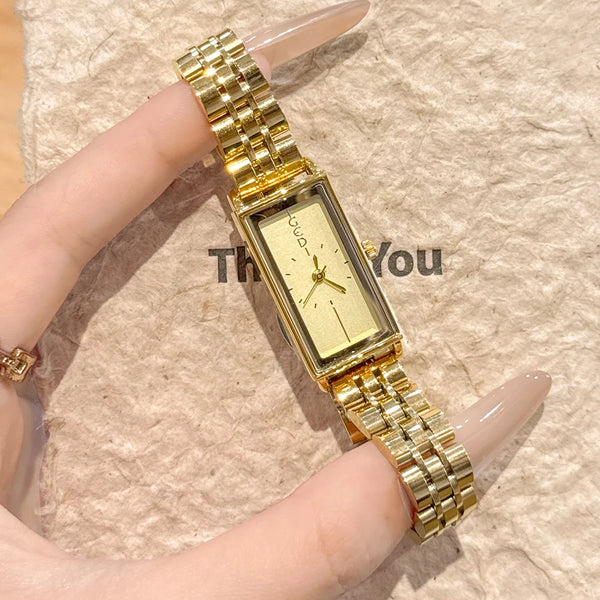 A close-up shot of a vintage gold stainless steel quartz watch worn on a finger against a beige background.