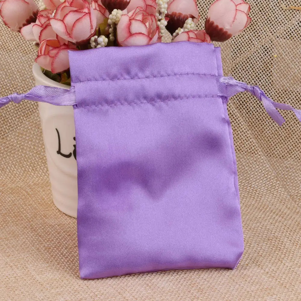 A purple satin drawstring jewelry pouch tied with a purple ribbon, resting on a beige fabric next to pink flowers.