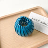 A close-up of a turquoise blue horsetail hair clip resting on a wooden tray with a white card.