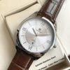 Montre argentée avec cadran blanc et bracelet en cuir marron, posée dans une boîte de couleur claire.