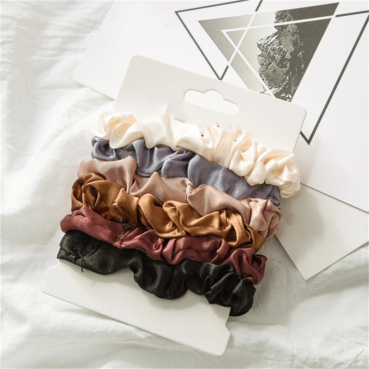 A pack of six satin hair scrunchies in various muted tones including beige, gray, mauve, and brown, displayed on a white card