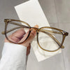 A hand holds a pair of large square anti-blue light spectacles with clear brown frames against a white background.