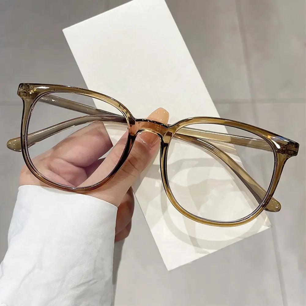 A hand holds a pair of large square anti-blue light spectacles with clear brown frames against a white background.