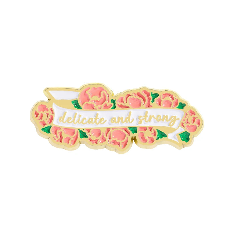 A gold enamel pin featuring a floral design with the text delicate and strong.