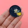 A hand holds a dark enamel pin featuring a cute firefly character on a black background.