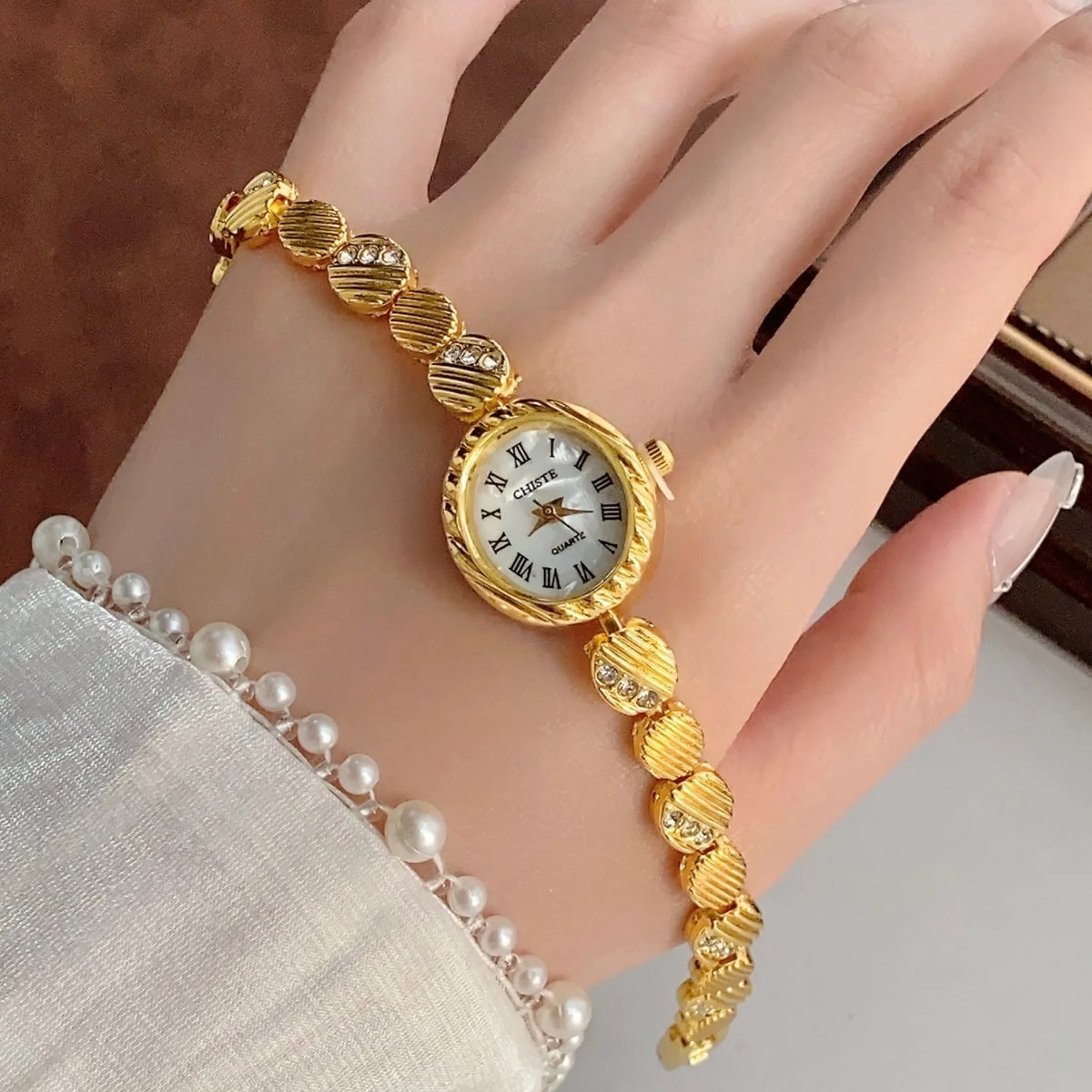 A gold-toned quartz fashion wristwatch with a round face and beaded bracelet worn on a hand.