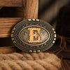 A vintage gold-toned belt buckle with an embossed letter E is displayed on a wooden surface.