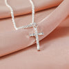 A delicate silver-plated cross necklace with sparkling CZ stones displayed on a pink fabric.