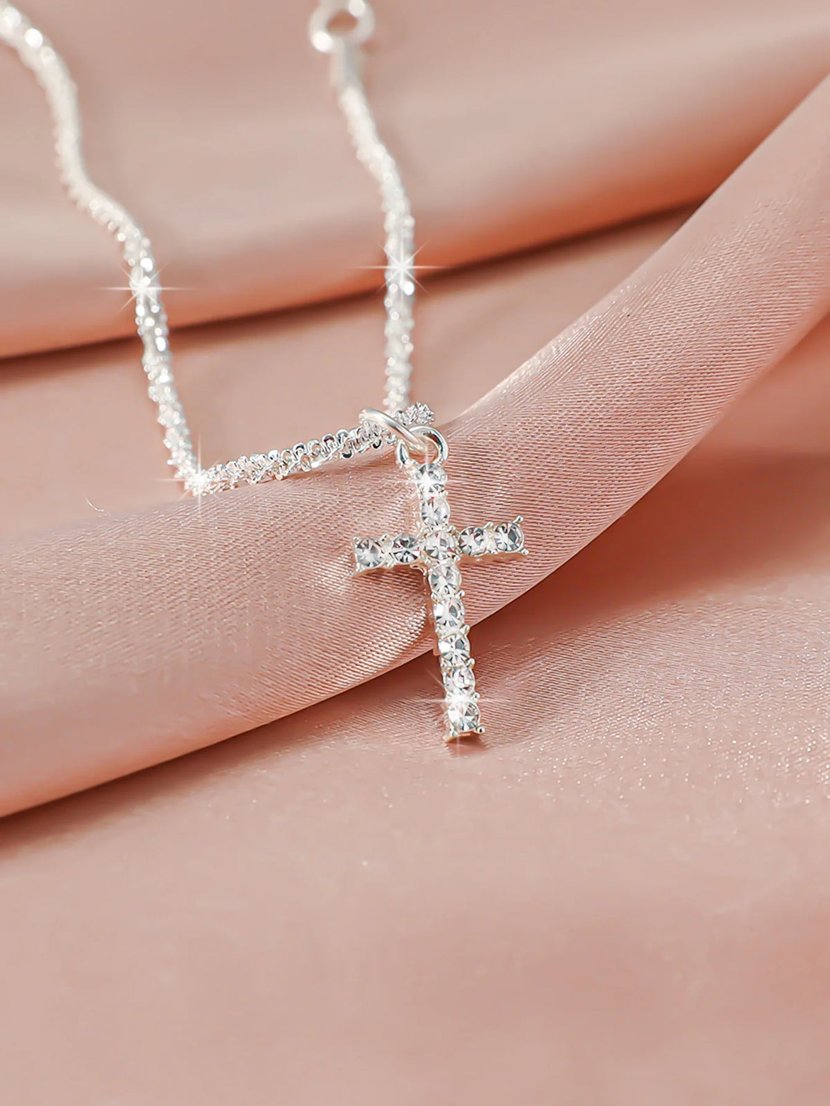 A delicate silver-plated cross necklace with sparkling CZ stones displayed on a pink fabric.