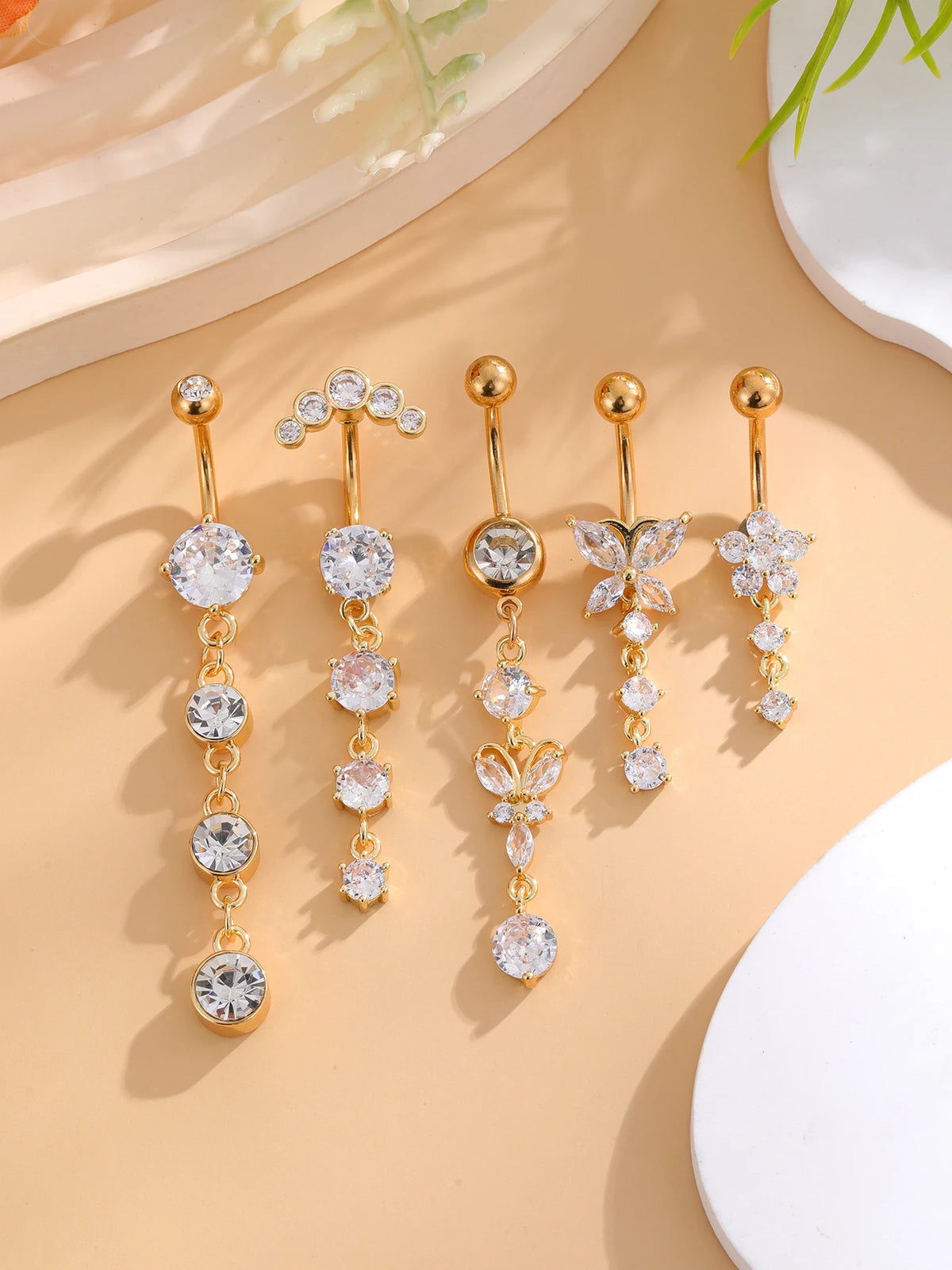 A set of five gold stainless steel dangled belly button rings featuring various diamond and pearl-like gemstone designs.