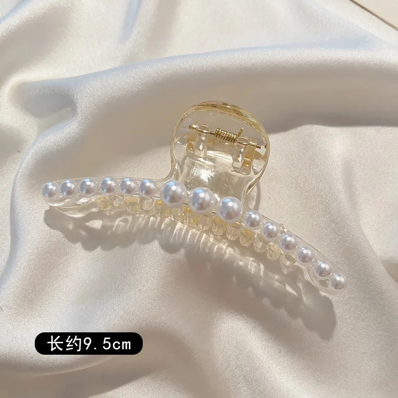 A clear plastic hair claw decorated with small white simulated pearls, shown against a white satin fabric.