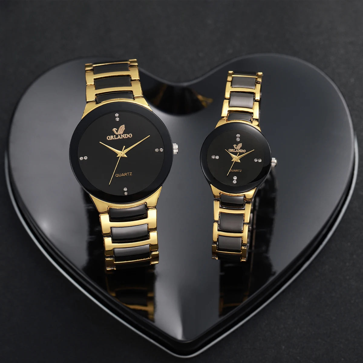 A pair of gold-toned analog quartz watches displayed on a black heart-shaped surface.