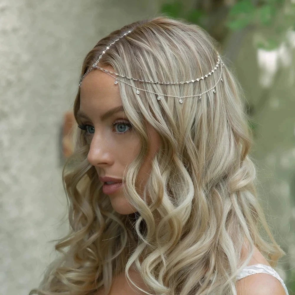 A woman with long blonde wavy hair wearing a delicate crystal head chain bridal hair accessory.