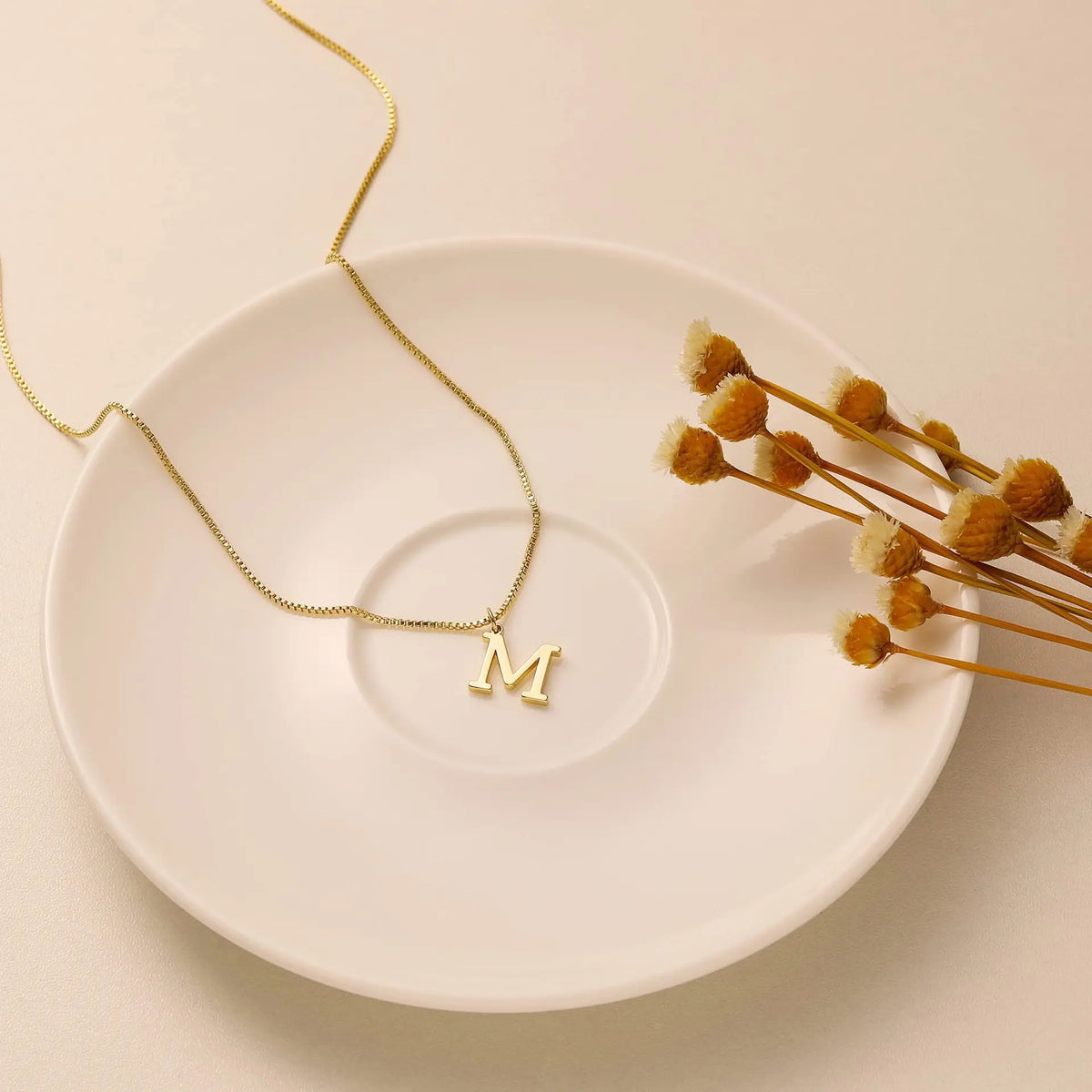 A delicate gold-plated initial letter necklace with a simple charm resting on a white ceramic plate.