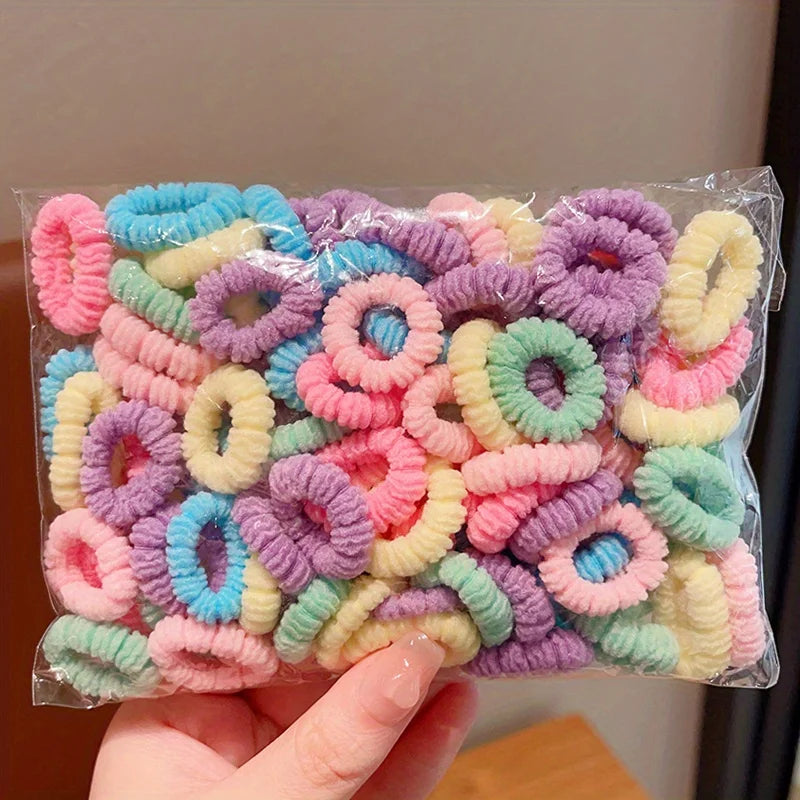 A hand holds a clear plastic bag filled with colorful elastic hair bands for girls in various shades of pink, purple, and yel