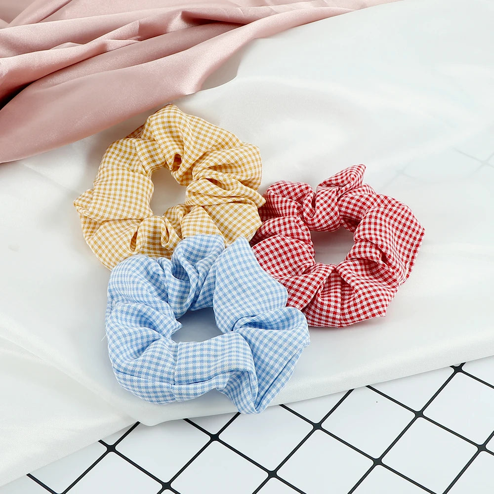 A flat lay of three colorful knitted hair scrunchie headbands in yellow, red plaid, and blue gingham on a white surface.
