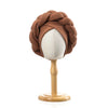 A brown braided turban cap displayed on a wooden mannequin stand.