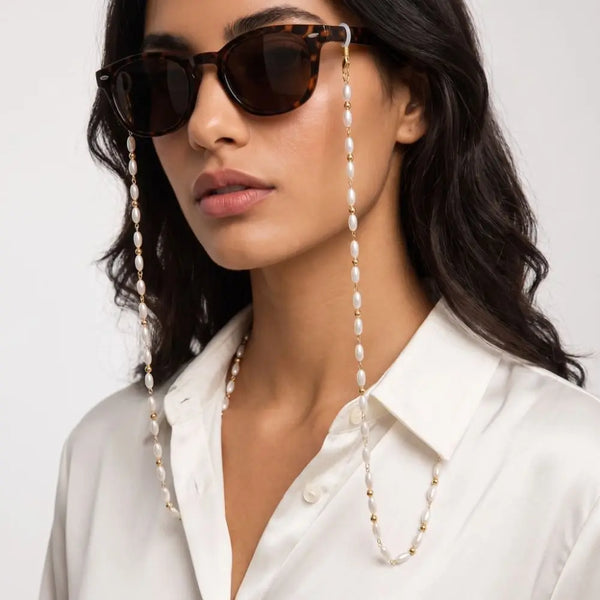 A woman wearing dark sunglasses with pearl and bead lanyards against a white collared shirt.