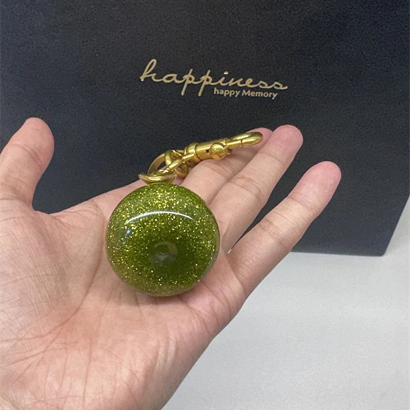 Une main tient un porte-clés de forme de poire verte cristalline avec une chaîne dorée sur fond sombre.