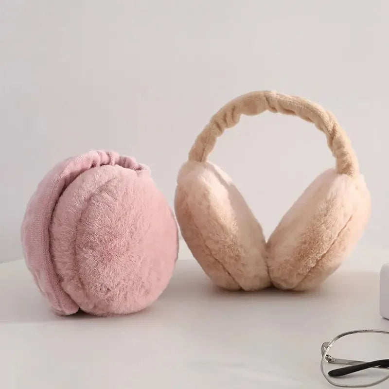A pair of light pink plush fur ear warmers or earmuffs with a woven headband shown on a white surface.