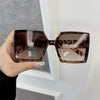A hand holds a pair of brown leopard print square sunglasses against a white door frame.