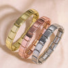 Three stacked stainless steel charm bracelets in gold, rose gold, and silver on a light background.