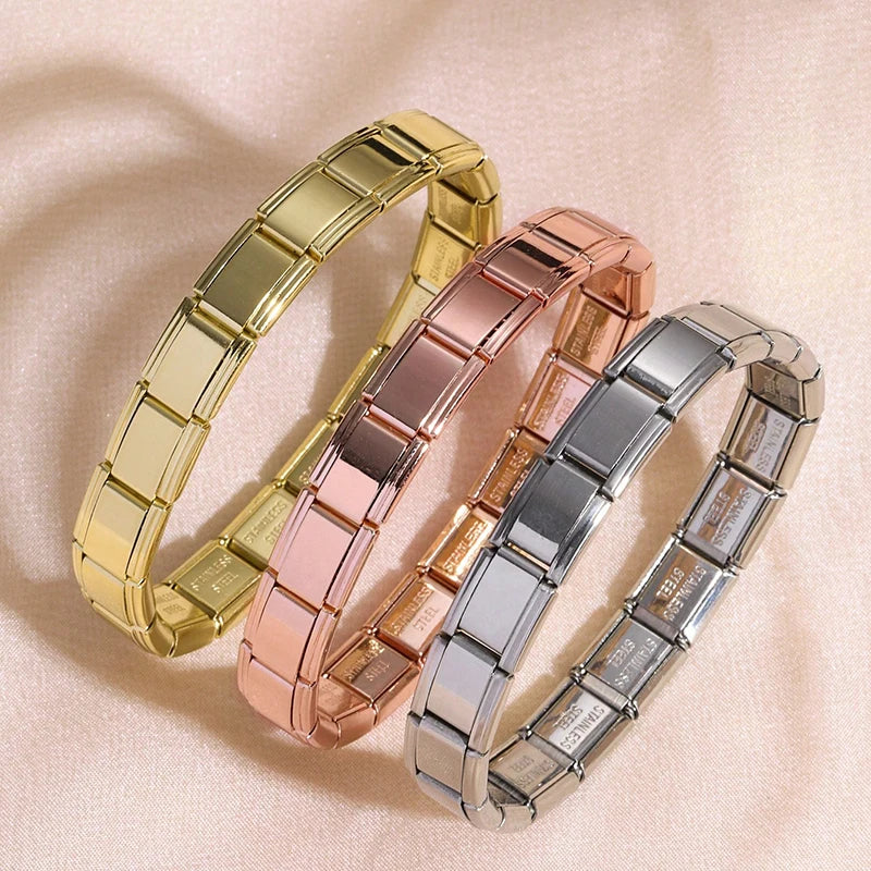 Three stacked stainless steel charm bracelets in gold, rose gold, and silver on a light background.