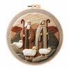 A circular embroidery design featuring a shepherd and sheep in earthy tones on a natural fabric background.
