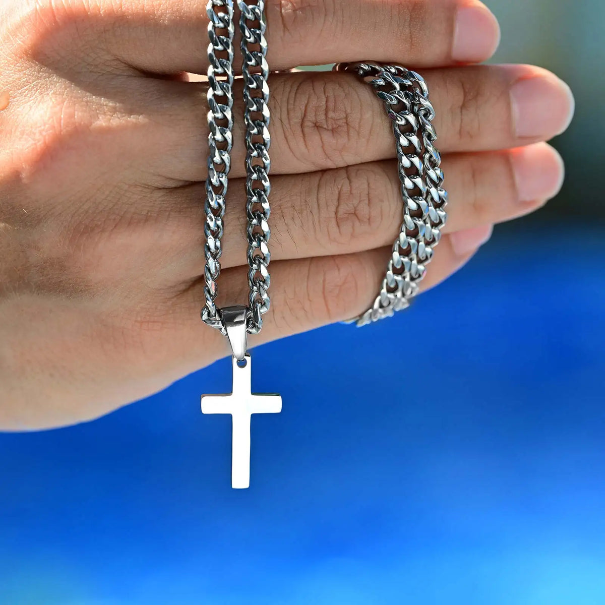 A hand is holding a stainless steel Cuban link necklace with a cross pendant.