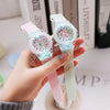 A hand holds a pink unicorn-themed fashion watch with a light green strap, next to some books.