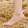 A close-up of a foot wearing gold plated initial letter anklets on golden sand.