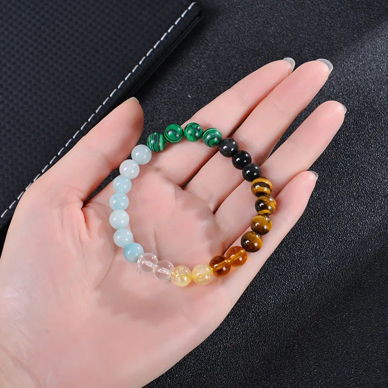 A hand holds a bracelet made of various colored tiger eye crystals, including green, black, yellow, and clear stones.