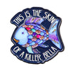 Broche émail bleu foncé en forme de poisson avec des écailles violettes et roses, affichant le texte "This is the skin of a k