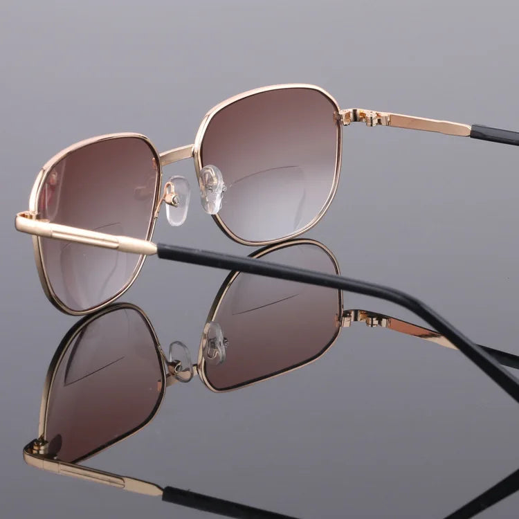 A pair of gold-toned sunglasses with rose-tinted lenses and clear bifocal inserts resting on a reflective surface.