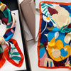 A floral print silk scarf featuring large flowers in shades of yellow, cream, and deep green against a dark background.