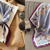 A light blue silk scarf with a floral print and purple trim is displayed on a beige garment.