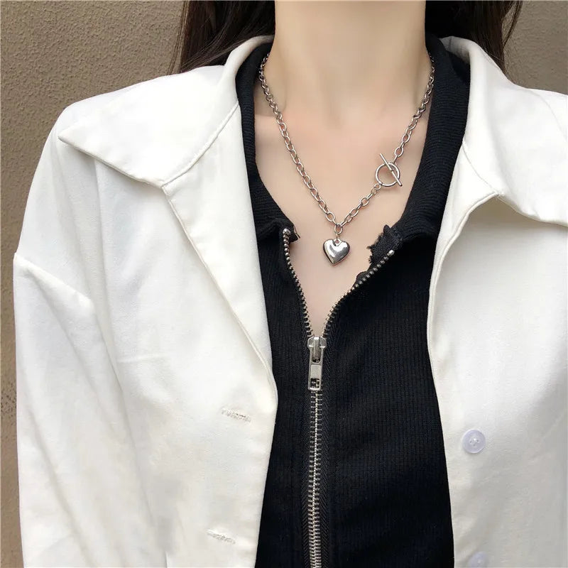 A close-up of a woman wearing a silver heart-shaped necklace under a black shirt and white jacket.