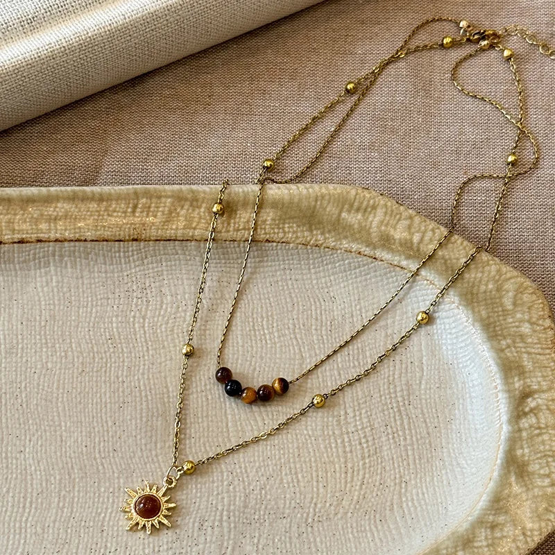 Gold chain necklace featuring a tiger eye sunflower pendant resting on a beige fabric surface.