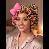 A woman wearing a leopard print hair drying cap with pink accents and a pink satin shirt.