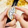 A hand is holding a gold-toned retro Y2K quartz watch with an irregular white face and blue hands against a blurred golden ba