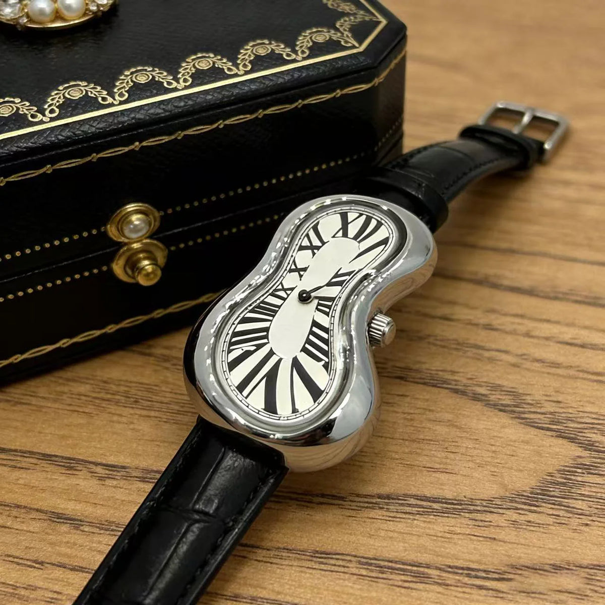 A silver irregular quartz watch with a black leather strap rests next to a black box.