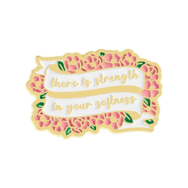 A gold enamel pin featuring a quote that reads there is strength in your softness surrounded by pink and green floral element