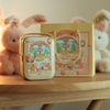 A decorative tinplate candy box with a gold carrying loop sits next to a fluffy pink rabbit toy.