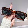 A hand holds a pair of tortoiseshell leopard print retro square sunglasses against a light background.